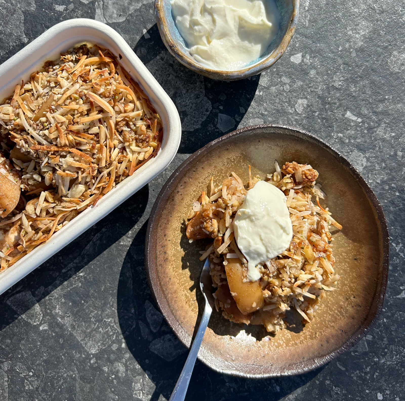 A serving of apple crumble with a dollop of cream on top, placed on a ceramic plate with a fork, next to a baking dish with more apple crumble.