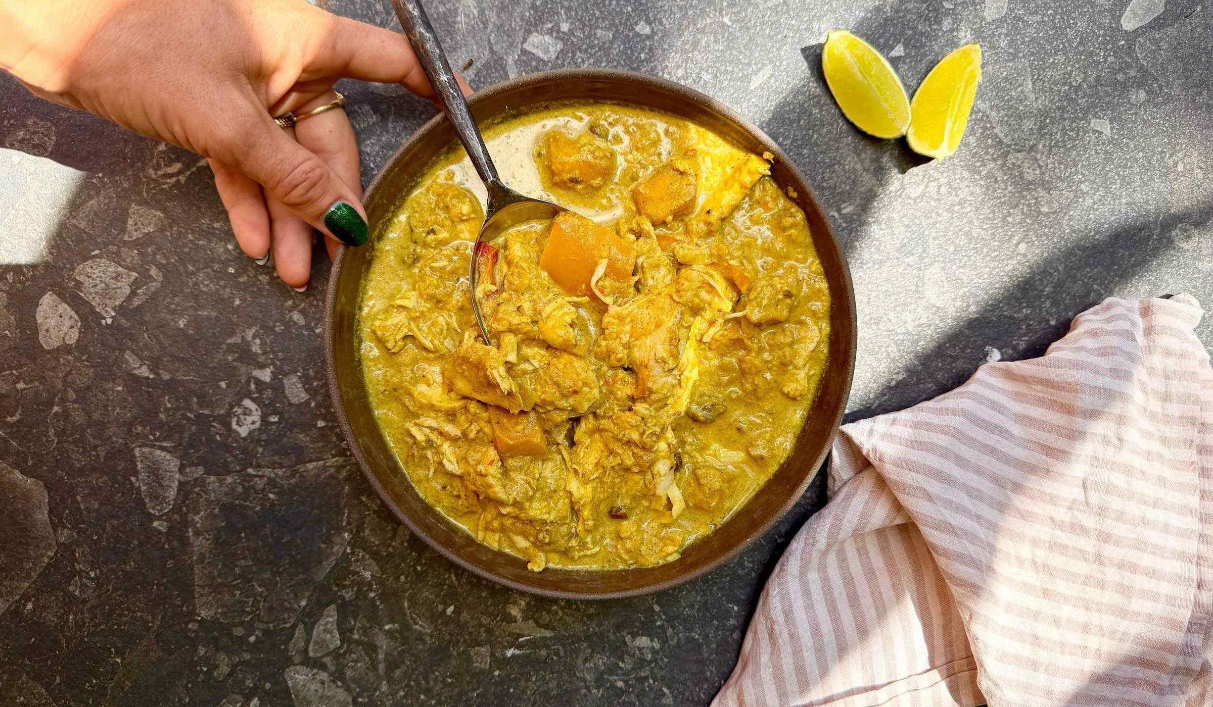 Bowl of yellow chicken mango curry with a spoon, on a dark surface with a striped cloth and lemon wedges.
