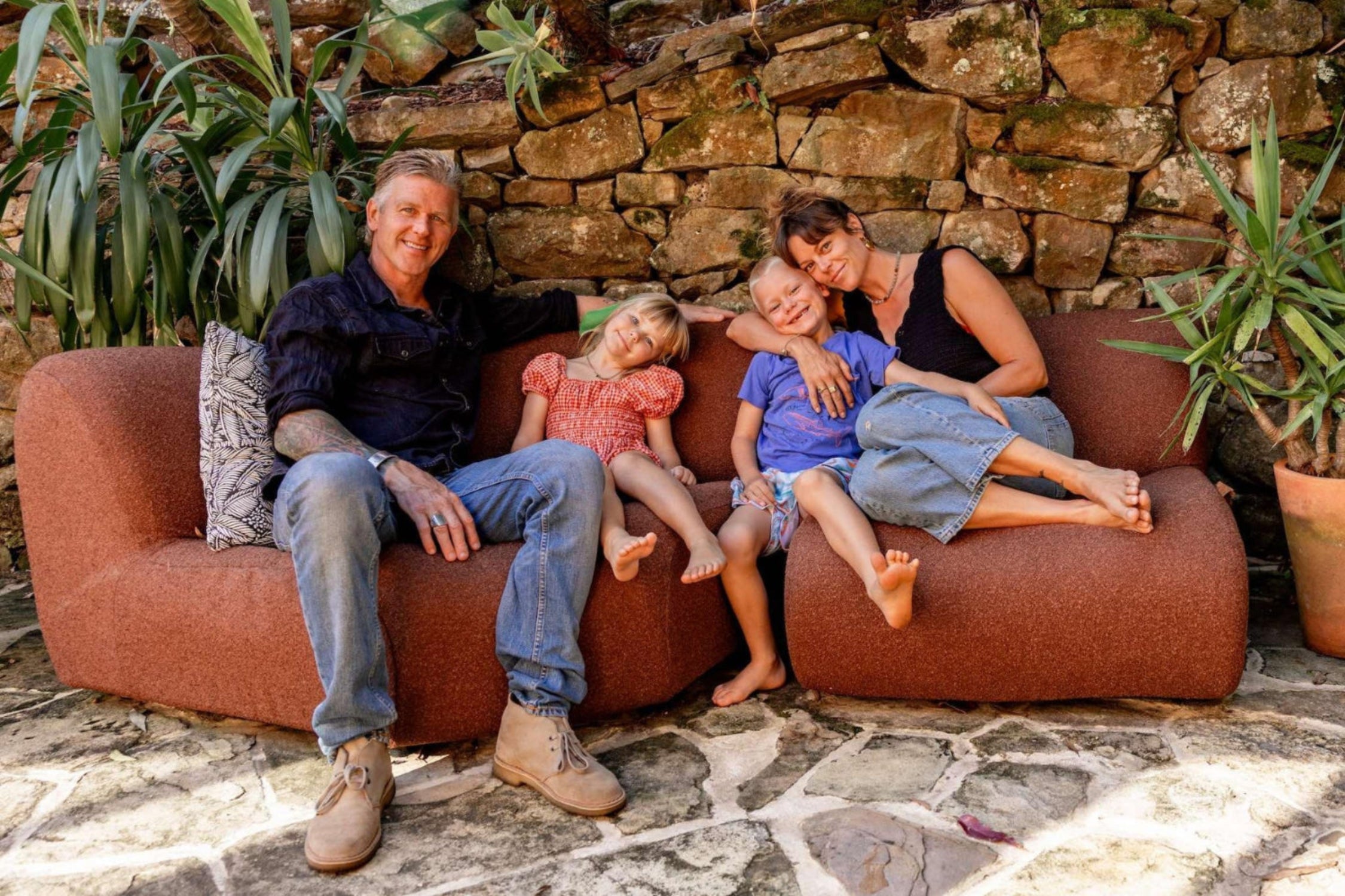 Family of four sitting on a red sofa against a stone wall with plants.