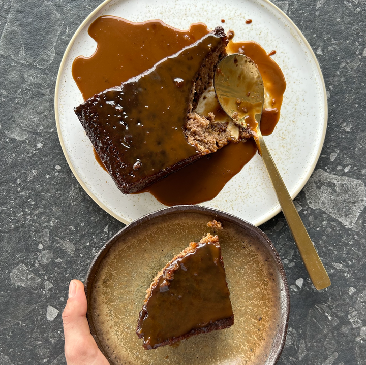 Hand holding slice of sticky date pudding with caramel sauce beside serving plate