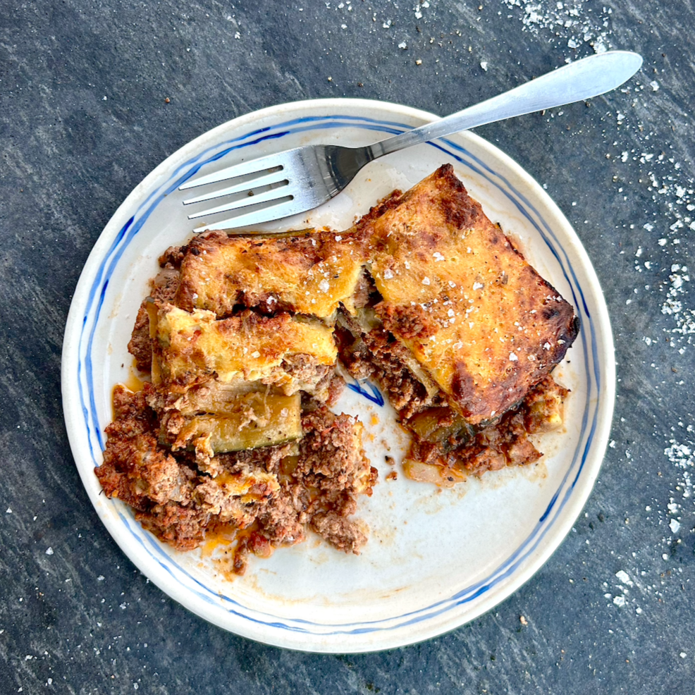 A plate with a serving of lamb moussaka, consisting of layers of eggplant and ground lamb, topped with béchamel sauce.