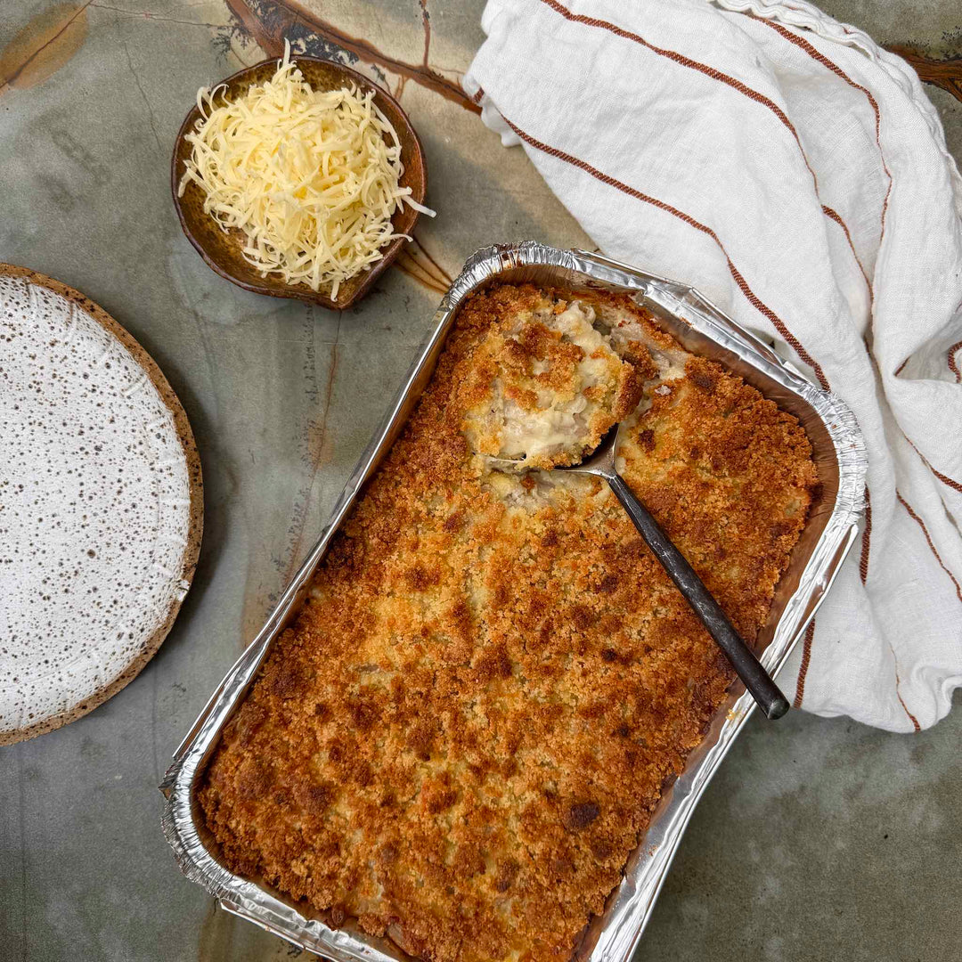 Baked dish in a foil pan with a spoon, next to a bowl of grated cheese and a speckled plate on a textured surface.