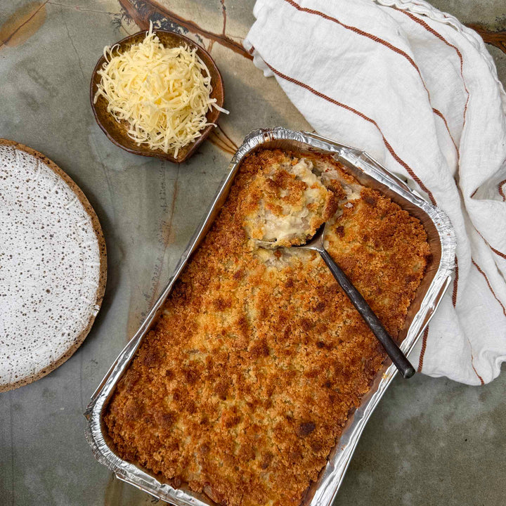 Baked dish in a foil pan with a spoon, next to a bowl of grated cheese and a speckled plate on a textured surface.