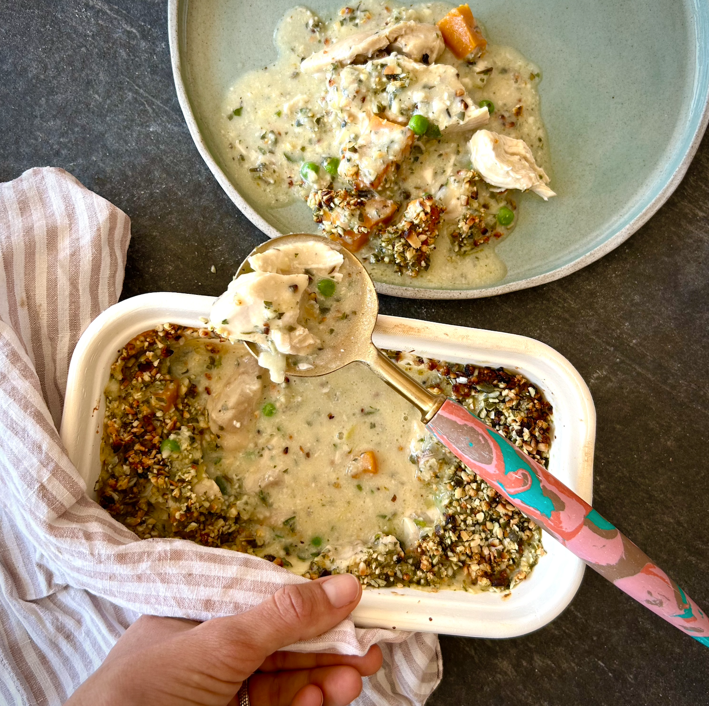 A hand holding a spoonful of organic chicken and leek pie, with a plate of the same pie in the background.