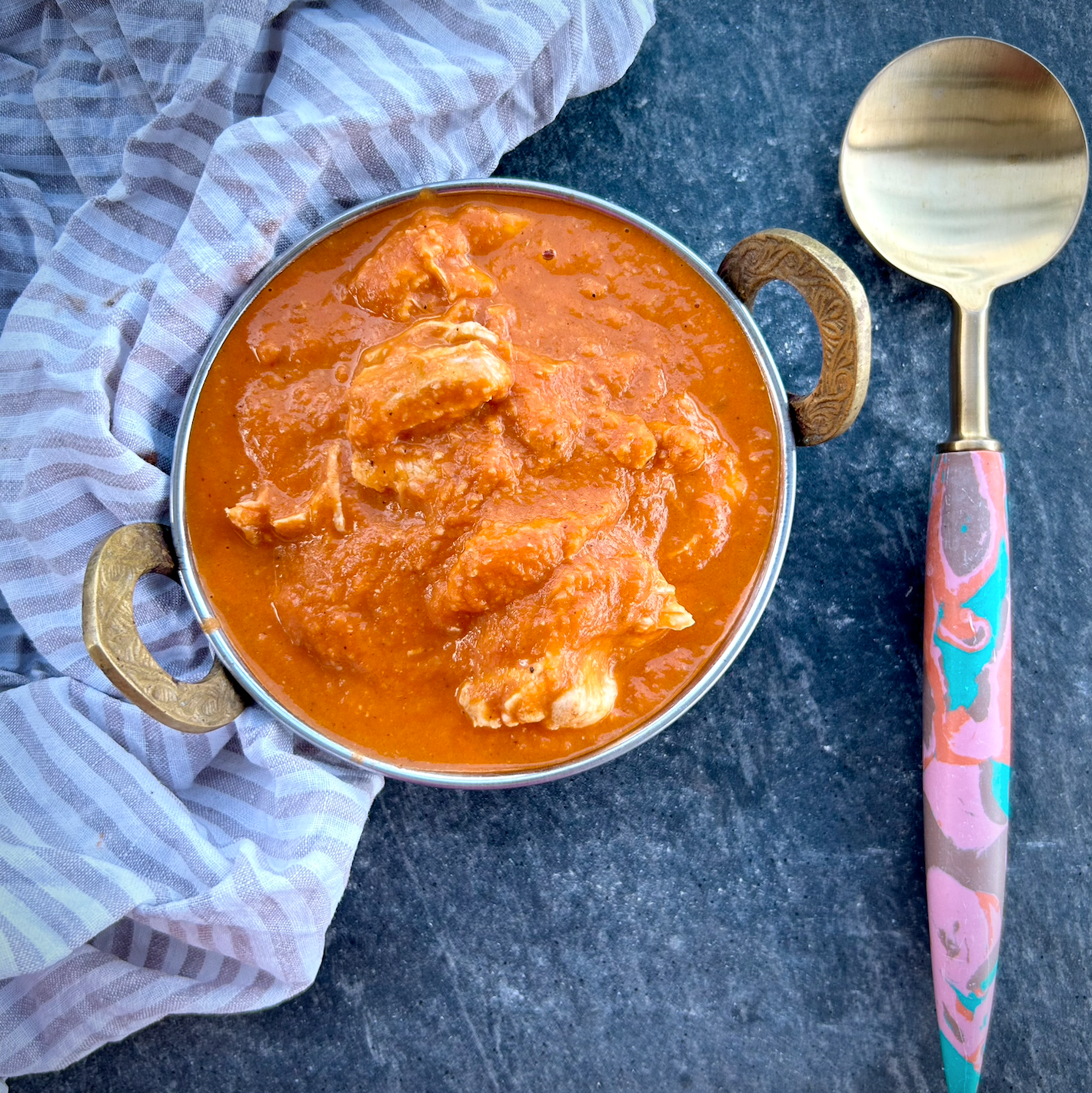 A bowl of butter chicken curry with a spoon on the side, placed on a striped cloth.