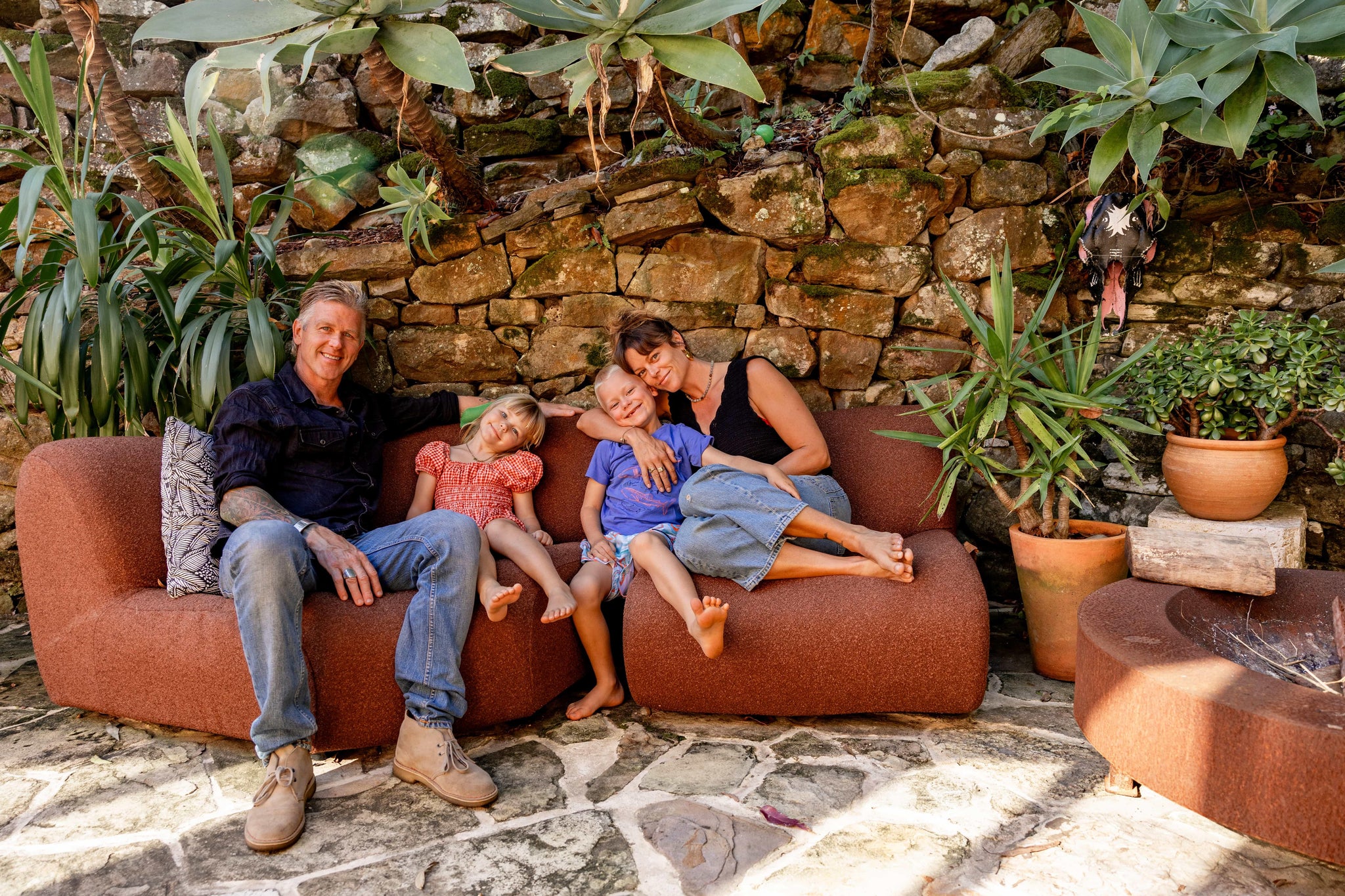Family relaxing on outdoor sofa with stone wall and potted plants in natural setting