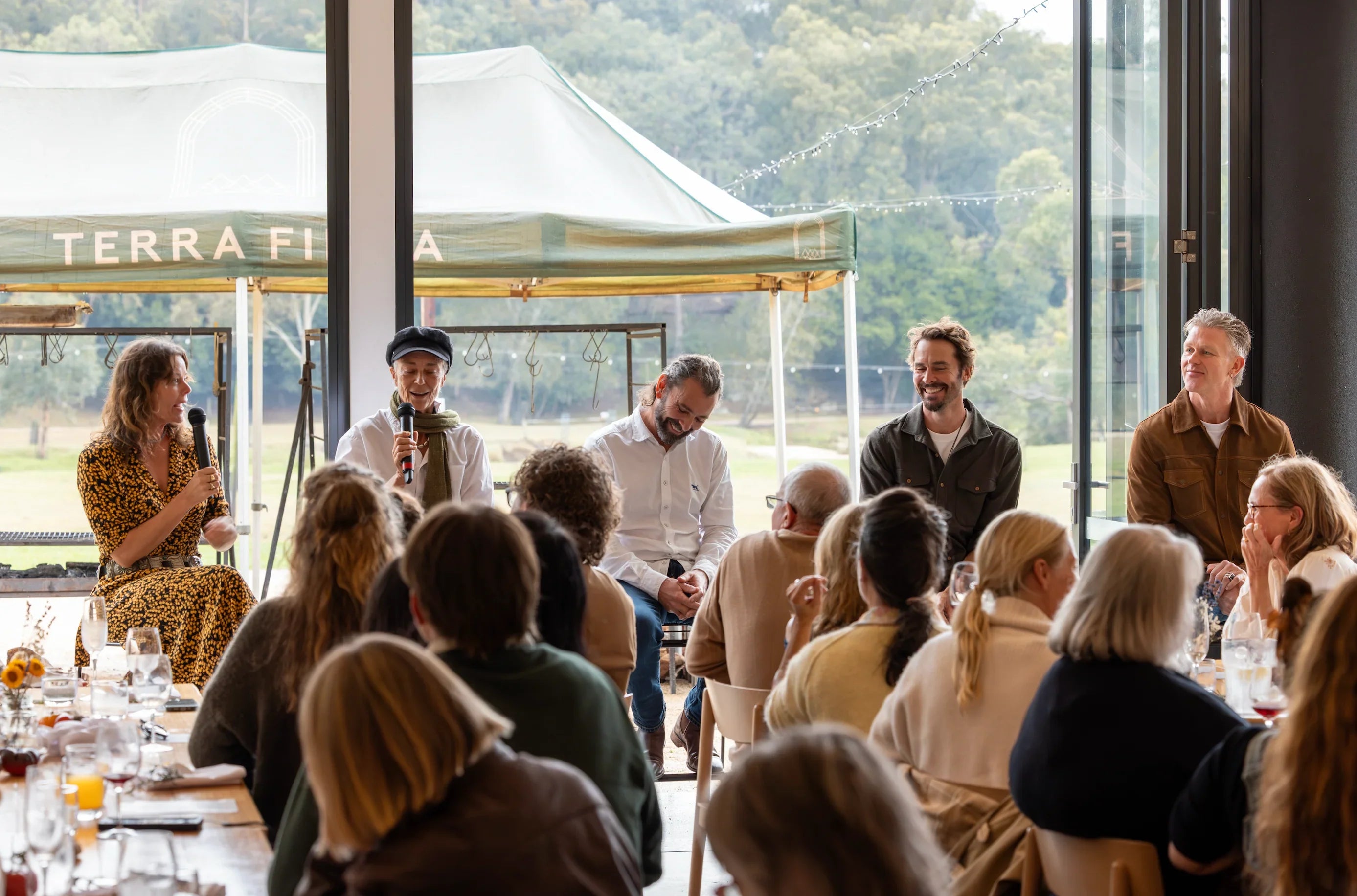 Group of people seated at a table with a speaker in front, outdoors with a tent labeled 'Terra Firma' in the background.